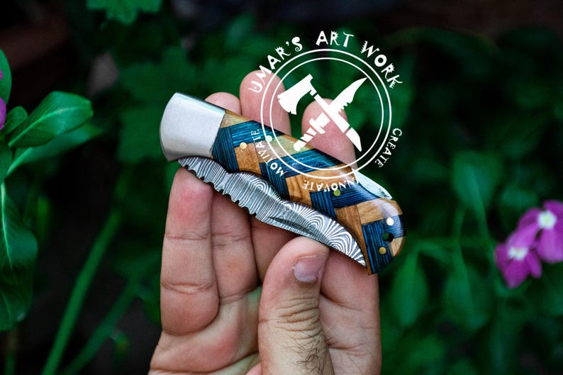 May include: A pocket knife with a blue and brown wood handle and a stainless steel blade. The blade has a wavy pattern and a serrated edge. The knife is closed and being held in a hand. The knife has the words "UMAR'S ART WORK CREATE INNOVATE" on the handle.
