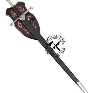 May include: A decorative sword with a silver blade and handle, and a black scabbard. The sword is mounted on a dark brown wooden plaque with gold-colored decorative accents. The plaque has a shield shape. The text "UMAR'S ART WORK" is visible.
