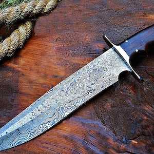 Puede incluir: Un cuchillo grande y ornamentado con una hoja estampada y un mango de madera oscura. La hoja tiene un dise&ntilde;o arremolinado y texturizado en tonos plateados y grises. El mango es marr&oacute;n oscuro con remaches plateados. El cuchillo se exhibe sobre una superficie de madera.