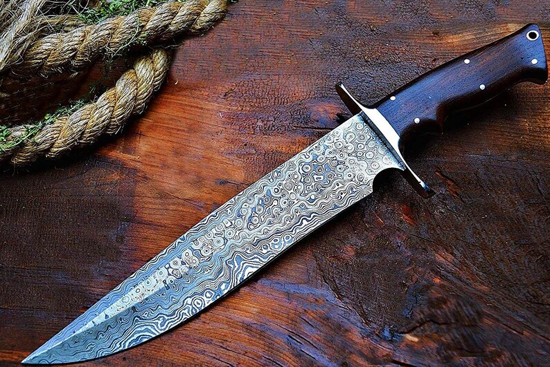 Puede incluir: Un cuchillo grande y ornamentado con una hoja estampada y un mango de madera oscura. La hoja tiene un dise&ntilde;o arremolinado y texturizado en tonos plateados y grises. El mango es marr&oacute;n oscuro con remaches plateados. El cuchillo se exhibe sobre una superficie de madera.