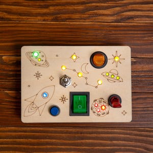 Può includere: Un pannello di legno con un design a tema spaziale, che presenta un razzo, pianeti, stelle e un UFO. Il pannello ha diversi pulsanti e interruttori, tra cui un interruttore a levetta verde, un pulsante rosso e un interruttore a levetta argentato.