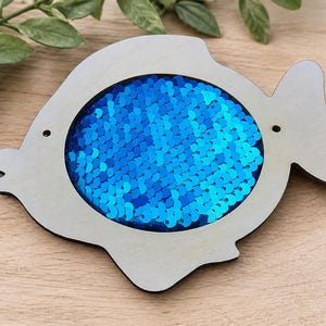 Pode incluir: Um brinquedo de madeira em forma de peixe com uma moldura cinza clara e um centro preenchido com lantejoulas azuis. O peixe tem uma boca recortada e dois pequenos buracos. O brinquedo está sobre uma superfície de madeira.