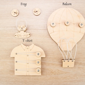 Peut inclure: Ensemble de jouets en bois avec une grenouille, un ballon à air chaud, un t-shirt et des boutons. La grenouille est assise sur un nénuphar et a une boucle pour l'accrocher. Le ballon à air chaud a trois boutons pour attacher le panier. Le t-shirt a quatre boutons pour attacher les manches.
