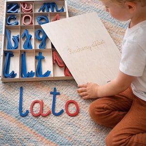 May include: A wooden box with compartments filled with blue and red wooden letters. The word "lato" is spelled out in blue and red letters. A child is holding a wooden board with the words "Ruchomy alfabet" engraved on it.