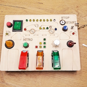 Op de afbeelding: Houten speelgoed raceauto dashboard met een rode, groene en gele schakelaar, een groene nitro-knop, een zilveren knop en een rood, geel en groen lichtpaneel. Het dashboard heeft ook een rode startknop, een blauwe stopknop en een sleutelgat. De tekst "STOP" en "START" staat op het dashboard gedrukt.