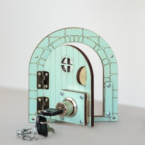 May include: A whimsical, light blue wooden door with an arched top, resembling a fairy door. It features a round window with a cross design, a key in the lock, and a set of keys. The door is decorated with faux stone detailing and metal hardware.