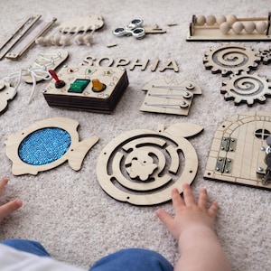 Könnte beinhalten: Ein hölzernes Activity-Board-Set mit verschiedenen interaktiven Elementen, darunter ein Zahnradsystem, ein Labyrinth, eine Tür mit Schloss und Schlüsseln, eine Schnürkomponente, eine Schalterbox, ein Perlen-Abakus und Holzbuchstaben für den Namen SOPHIA.