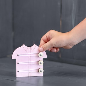 Può includere: Un giocattolo in legno a forma di camicia rosa chiaro con tre bottoni circolari in legno fissati con cordini elastici bianchi. Il giocattolo presenta un dettaglio del colletto e piccoli fori per infilare, utile per esercitare la motricità fine.
