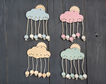 Puede incluir: Cuatro adornos de pared de madera con forma de nube, cada uno con un pequeño sol recortado en la parte superior. Cada nube tiene cinco cordones beige colgantes, con una cuenta de madera redonda al final de cada cordón. Las nubes están pintadas en azul claro, rosa, beige y verde menta.