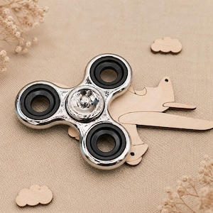 May include: A metallic silver fidget spinner with three black circular bearings positioned over a light-coloured wooden cutout of an aeroplane and two small wooden cloud shapes, all arranged on a textured beige fabric surface.