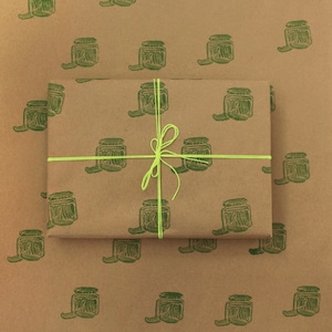 Puede incluir: Papel de regalo marrón con un patrón repetido de tarros de pepinillos verdes. Un trozo de papel de regalo más pequeño está encima del más grande, atado con una cinta verde brillante.