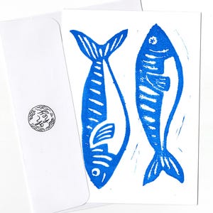 May include: A white card with two blue fish printed on it. The fish are stylised and have a simple design. The card is blank and ready for a message.