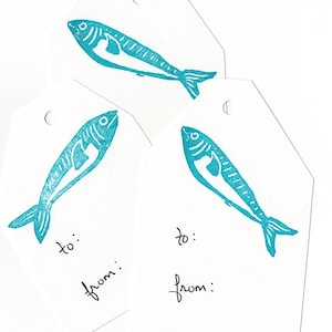 Puede incluir: Tres etiquetas de regalo blancas con un diseño de pez azul turquesa. Cada etiqueta tiene las palabras "para:" y "de:" escritas con tinta negra.