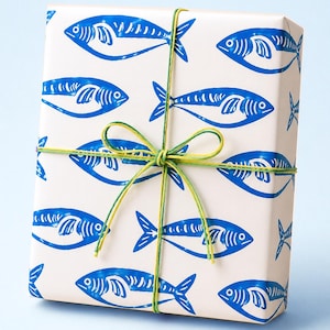 May include: A wrapped gift box featuring a repeating pattern of blue fish on a cream background. The box is tied with green and yellow twine, creating a nautical theme. The gift wrap is suitable for birthdays or other special occasions.