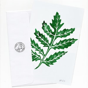 May include: A white greeting card featuring a green leaf print and a matching envelope. The leaf design has white veins, and the envelope has a small, circular stamp. The card's design is simple and inspired by nature.