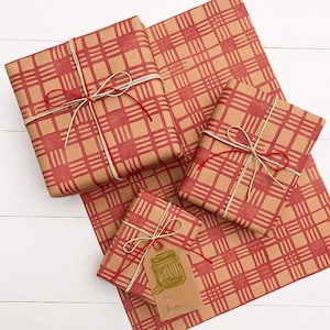 Puede incluir: Regalos envueltos con papel a cuadros rojo y cordel rojo y dorado. Una pequeña etiqueta de regalo con una ilustración de un tarro está adjunta a un paquete. Los regalos están dispuestos sobre una superficie de madera blanca.