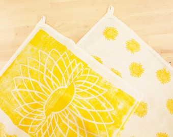 Op de afbeelding: Twee witte en gele keukendoeken. Eén doek heeft een groot geel zonnebloemontwerp, terwijl de andere meerdere kleinere gele zonnebloemprints heeft. Beide doeken hebben een lus om op te hangen.