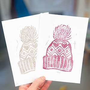 Puede incluir: Dos tarjetas con imágenes impresas a mano de gorros de invierno. Un gorro es plateado y el otro es rojo.