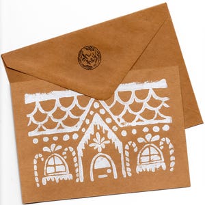 Può includere: Un set di biglietti di auguri e buste marroni. Il biglietto presenta un disegno di casa di pan di zenzero bianco disegnato a mano. La busta ha un timbro circolare nell'angolo in alto a sinistra. La carta è strutturata.