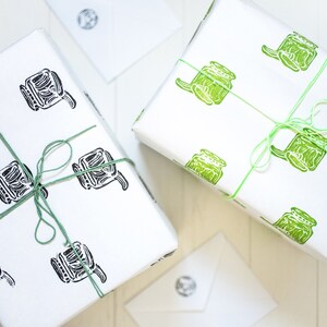Puede incluir: Dos cajas de regalo envueltas en papel blanco con un patrón repetido de botes verdes y negros. Las cajas están atadas con una cuerda verde.