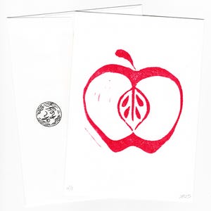 May include: A white greeting card featuring a red apple linocut print. The apple design includes a leaf detail. The card is folded, with a small black and white circular design on the left. The card is numbered 4/5.