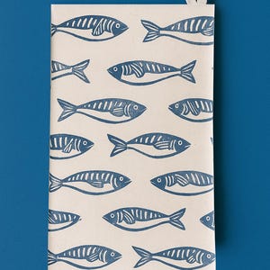 May include: A cream-colored notebook with a repeating pattern of blue fish illustrations. The fish are stylized with detailed fins and scales. A small loop is at the top for hanging. The background is a solid blue.