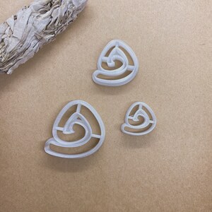 May include: Three white plastic cookie cutters in a spiral design. The cutters are in three different sizes, with a sage bundle in the upper left corner. The cutters are on a tan surface.