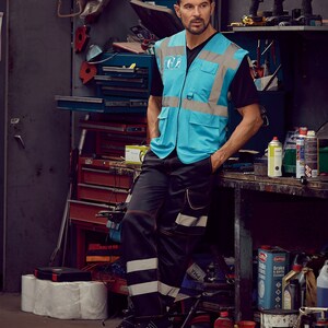 May include: A man wearing a blue safety vest with reflective strips and black work trousers with reflective stripes. He is standing in a garage setting with tools and supplies in the background.