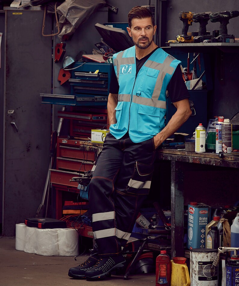 May include: A man wearing a blue safety vest with reflective strips and black work trousers with reflective stripes. He is standing in a garage setting with tools and supplies in the background.