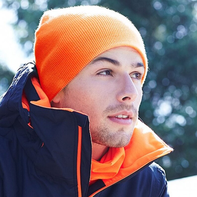 May include: A bright orange beanie hat with a ribbed texture. The hat is worn by a person wearing a dark blue jacket with an orange fleece lining.