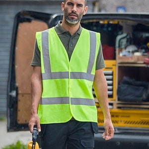 May include: A man wearing a green polo shirt and a bright yellow safety vest with reflective silver stripes. He is holding a pair of yellow earmuffs.