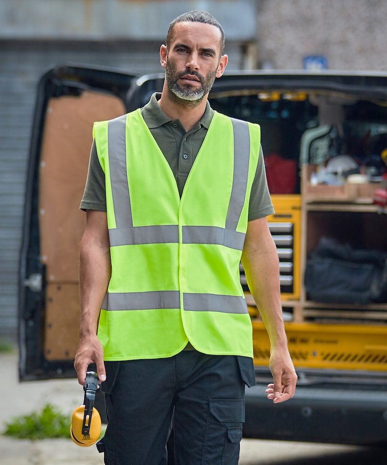 May include: A man wearing a green polo shirt and a bright yellow safety vest with reflective silver stripes. He is holding a pair of yellow earmuffs.