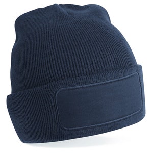 Puede incluir: Un gorro azul marino con un gran parche rectangular en blanco en la parte delantera. El gorro est&aacute; hecho de un tejido suave de punto.