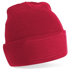 Puede incluir: Un gorro de punto rojo con un parche rectangular en blanco en la parte delantera.