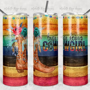 Può includere: Un bicchiere in acciaio inossidabile con un design a venature di legno colorato. Il bicchiere presenta un paio di stivali da cowboy marroni con dettagli floreali e il testo "Busy doing cowgirl stuff".
