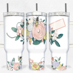 Puede incluir: Un vaso de acero inoxidable blanco con un diseño floral y una abeja. El vaso tiene una tapa y un asa. Las flores son rosas, amarillas y moradas. La abeja es amarilla y negra.