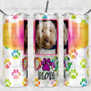 May include: A white tumbler with a colorful watercolor design and paw prints. The tumbler has a photo of a dog in the center and the words "Dog Mom" in glitter letters.