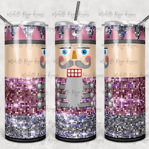 May include: A tumbler featuring a nutcracker design. The tumbler has a light tan face with blue eyes, a gray mustache, and a red mouth. The top and bottom of the tumbler are decorated with pink and silver glitter. A silver straw is included.
