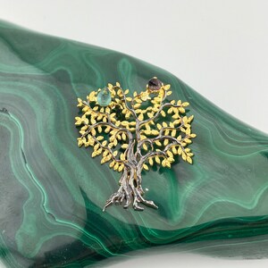 May include: A gold and silver tree of life pendant with green and purple gemstones. The pendant is on a green malachite background.