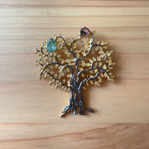 May include: A gold-toned tree of life brooch with a light blue and pink gemstone accent. The tree is intricately detailed with leaves and branches.