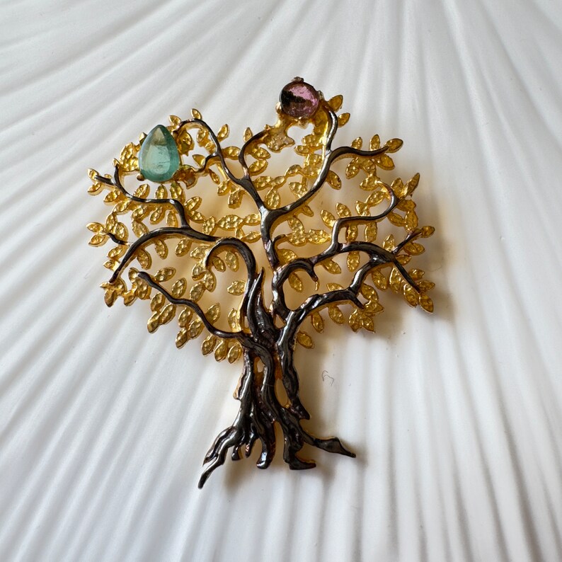 May include: A gold and black tree of life brooch with pink and green gemstones.