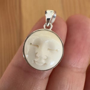 May include: A round pendant featuring a carved off-white face with closed eyes and a subtle smile, set within a polished silver frame. The pendant includes a silver bail at the top for attachment to a chain, showcasing a smooth, glossy finish.