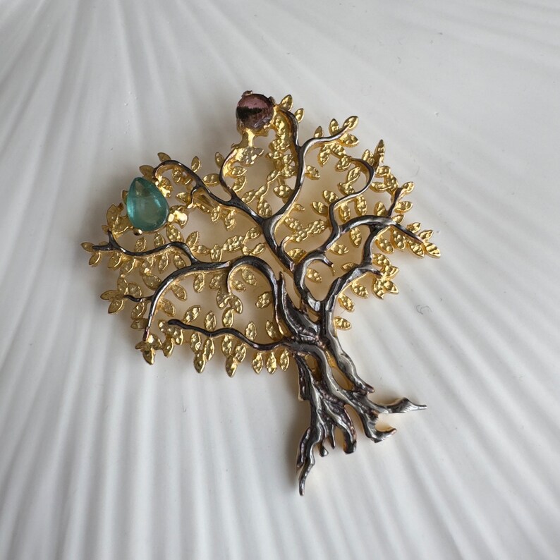 May include: A gold and silver tree of life pendant with green and pink gemstones. The pendant features a detailed design with intricate branches and leaves.
