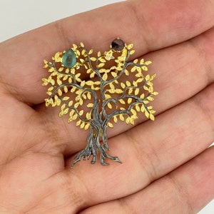 May include: A gold and black tree of life pendant with three gemstones. The tree is detailed with leaves and branches. The gemstones are green, red, and black.