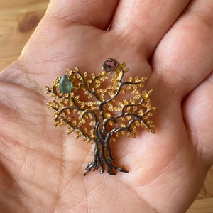 May include: A gold and black tree of life pendant with green and pink gemstones. The pendant is on a hand.