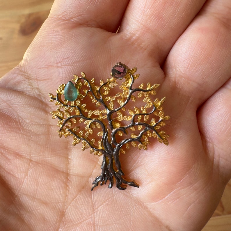May include: A gold and black tree of life pendant with green and pink gemstones. The pendant is on a hand.