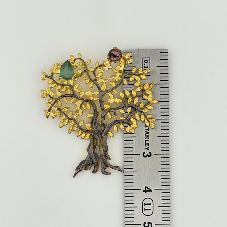 May include: A gold and black tree of life pendant with green and pink gemstones. The pendant is approximately 2 inches tall.