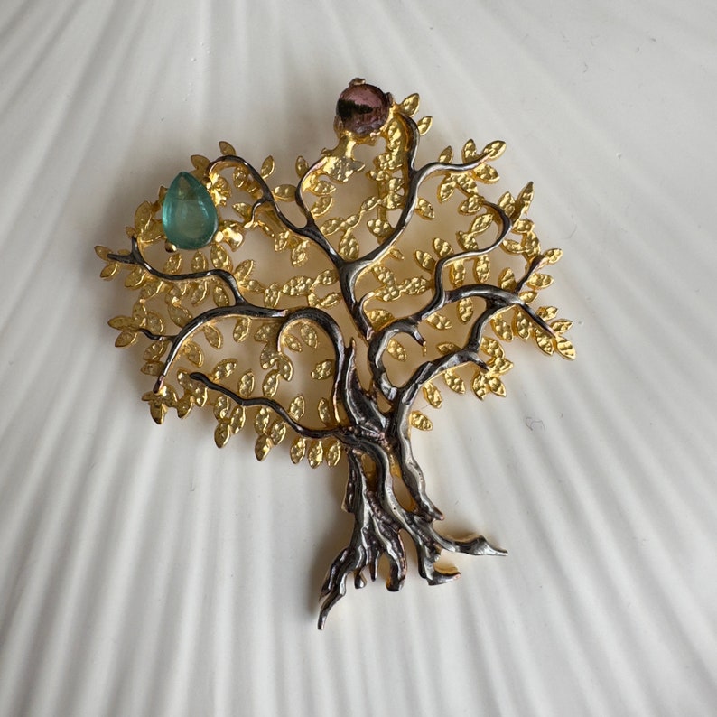 May include: A gold and silver tree of life pendant with green and pink gemstones. The pendant is detailed with intricate branches and leaves.