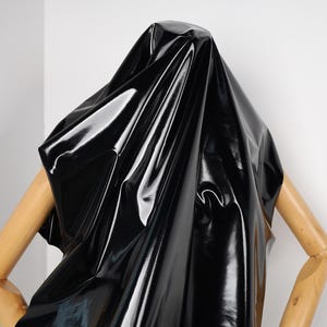 Peut inclure: Un tissu noir brillant recouvre la tête et les épaules d'un mannequin. Le tissu reflète la lumière, créant des reflets et des ombres. Les bras en bois du mannequin sont visibles, pliés aux coudes.