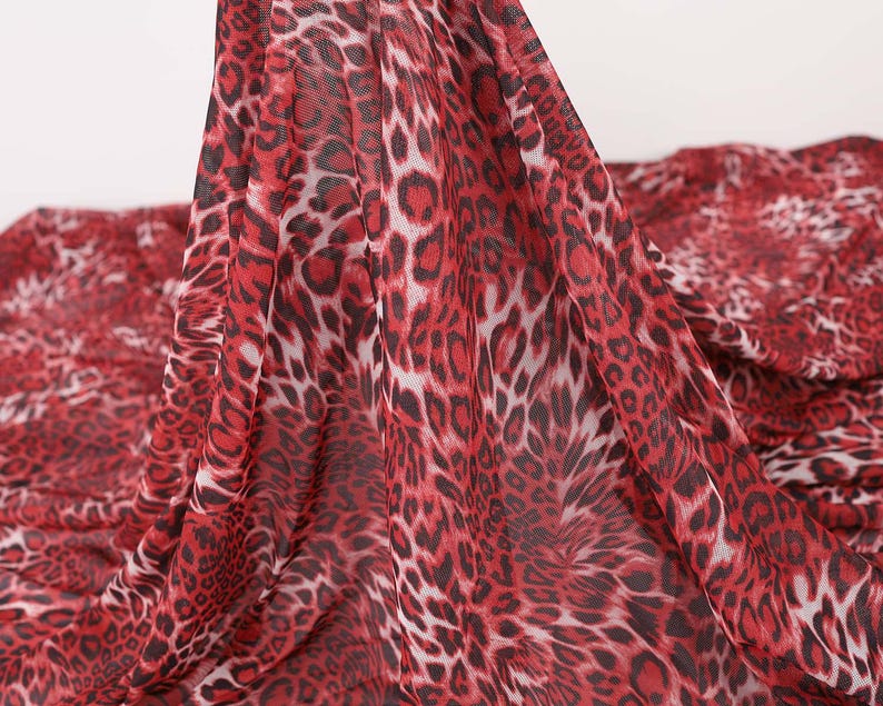 May include: Red and black leopard print fabric. The fabric has a sheer, lightweight appearance. The leopard spots are a deep red color with black outlines and white centers. The fabric is draped, showing the pattern's flow.